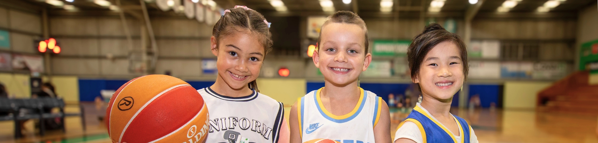 Three children holding basketballs smile at the camera while standing on a basketball court